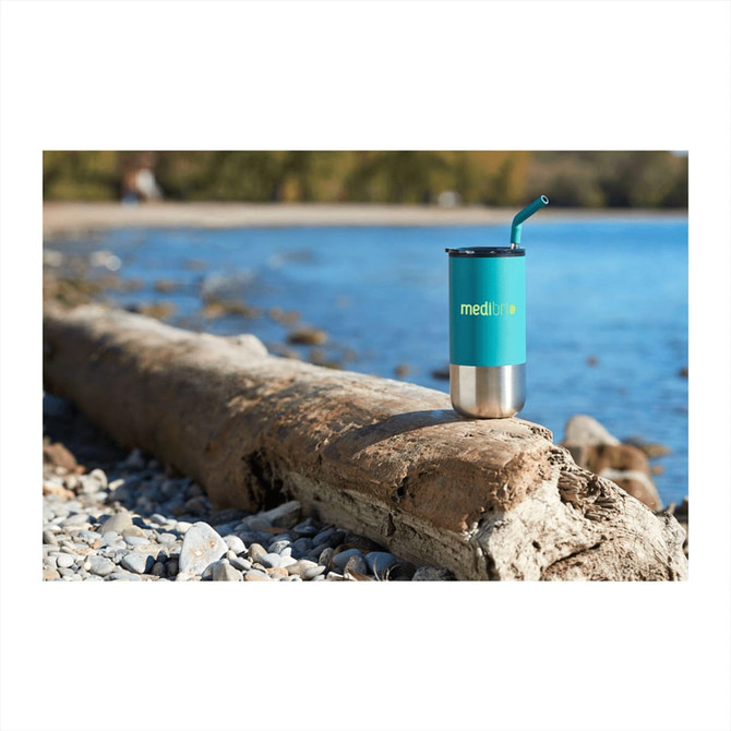 A drink bottle, turquoise with a stainless steel base and straw, placed on a log by the water's edge.