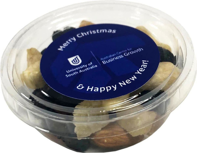 A clear tub containing a mix of nuts and dried fruit, featuring a blue lid with a logo and festive message.