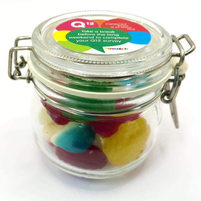 A glass canister filled with colourful mixed lollies, featuring green, red, yellow, and white sweets, with a branded lid.