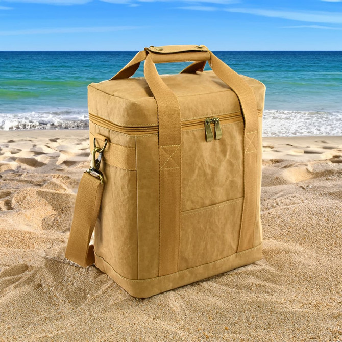 A tan Kraft paper lunch cooler bag with handles and a shoulder strap, placed on sandy beach terrain by the sea.