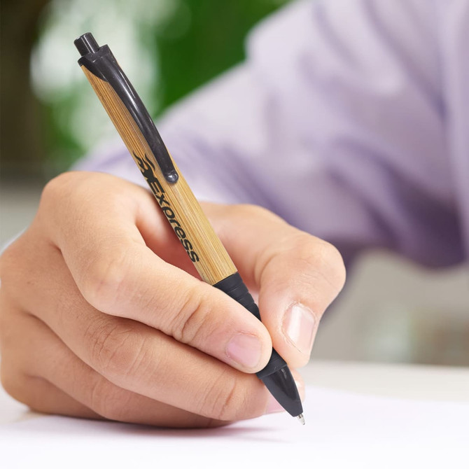 A bamboo and black plastic pen held in a hand, writing on a white surface, with a logo visible on the pen.