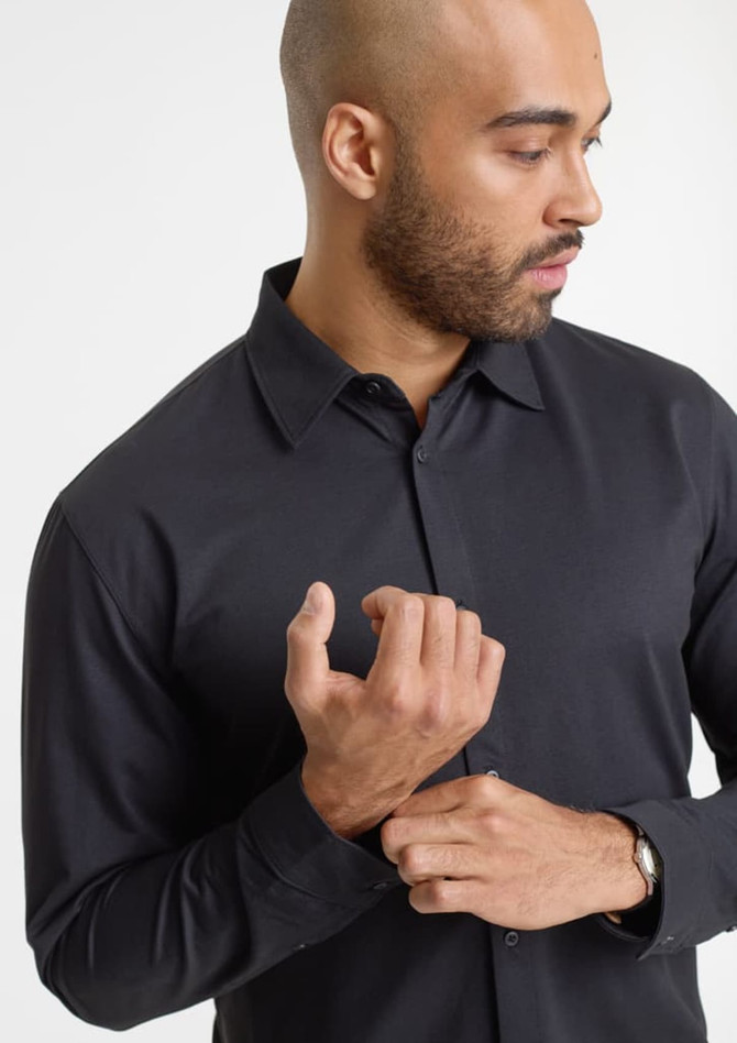 A black long sleeve shirt on a model, featuring a collar and button-up design. The model is adjusting his shirt.