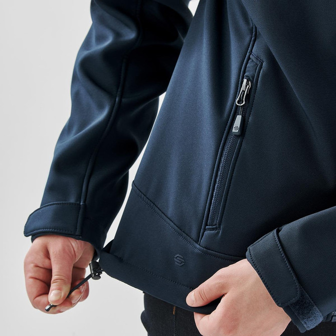 A close-up of a women's navy softshell jacket featuring a zipped pocket and adjustable hem. The jacket has a brand logo.