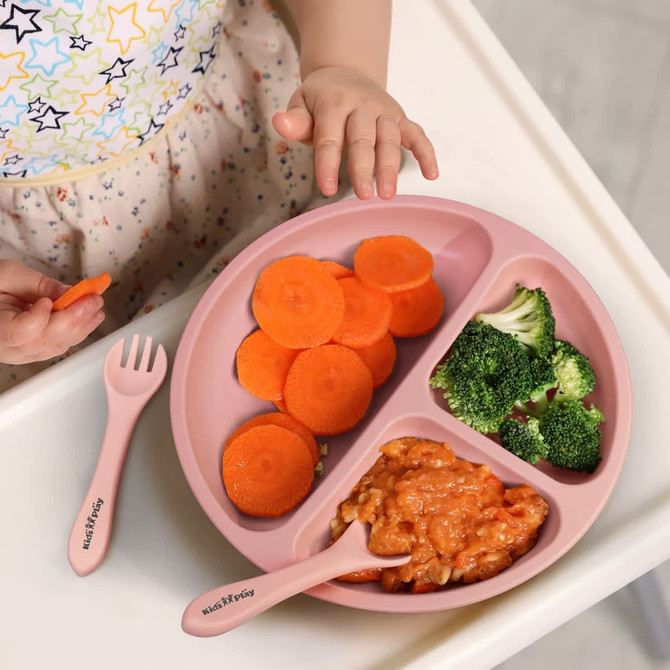 A pink kids' plate set with compartments holds sliced carrots, broccoli, and sauce-covered pasta; accompanied by a fork and spoon.