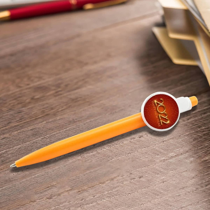 An orange plastic pen with a circular badge on the side, resting on a wooden surface beside stationery.