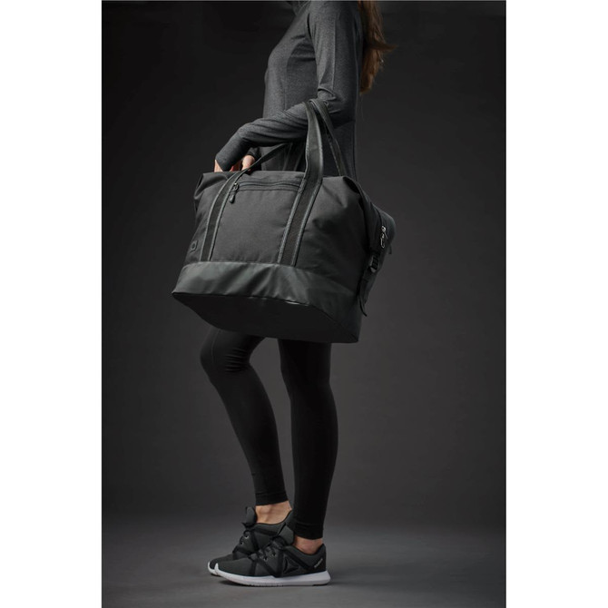 A duffle bag in black being held by a person wearing athletic attire and sneakers. The bag features a logo.