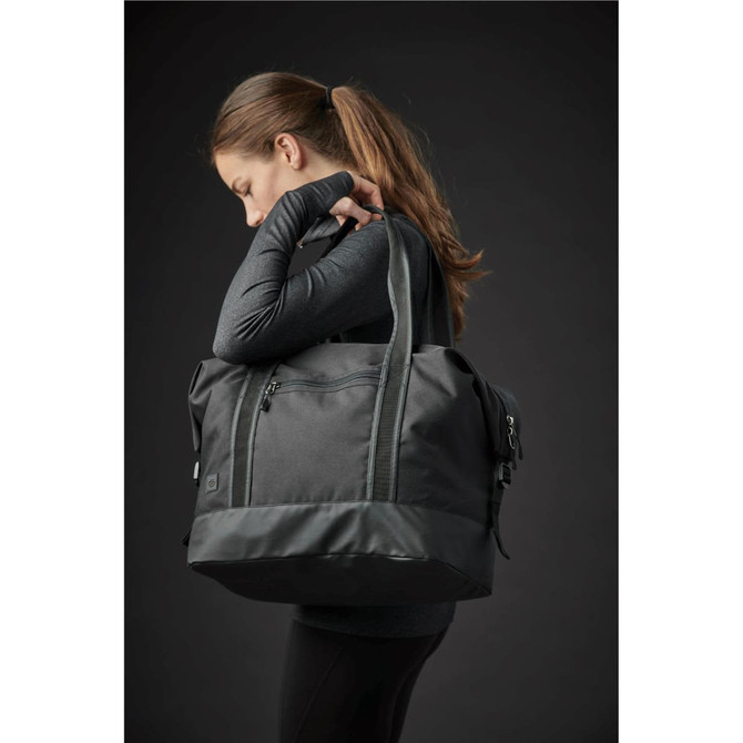 A black duffle bag with a logo, carried by a person, featuring dual handles and zippered compartments.