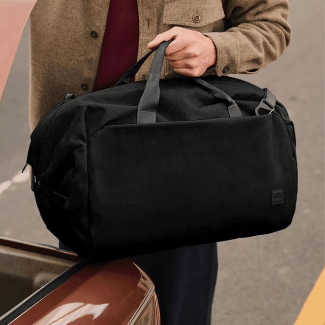 A black duffel bag with carrying handles is held by a person near a car trunk. The bag has a logo on it.