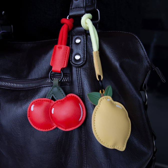 A black handbag with two bag charms: a red cherry pair and a yellow lemon, featuring green and yellow accents.