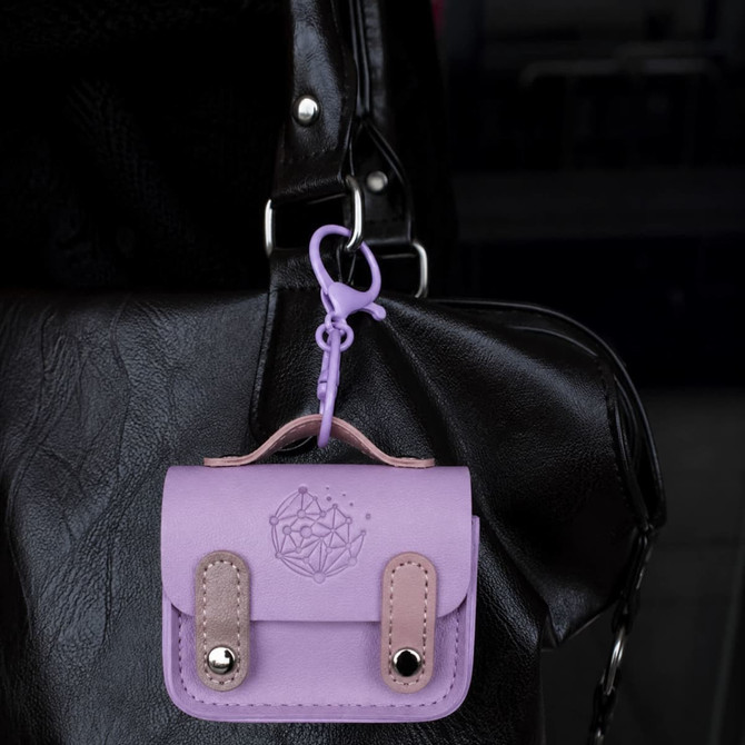 A small, lavender mini tech charm case with a clasp hanging from a black handbag. It has a decorative design and two tan straps.