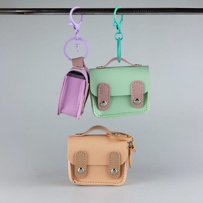 Three small tech charm cases in pink, green, and peach hanging from a display, featuring various snap and zipper closures.