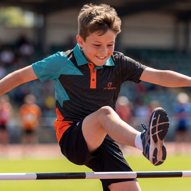 A boy jumps over a high jump bar while wearing a custom active polo in black, teal, and orange with a logo.