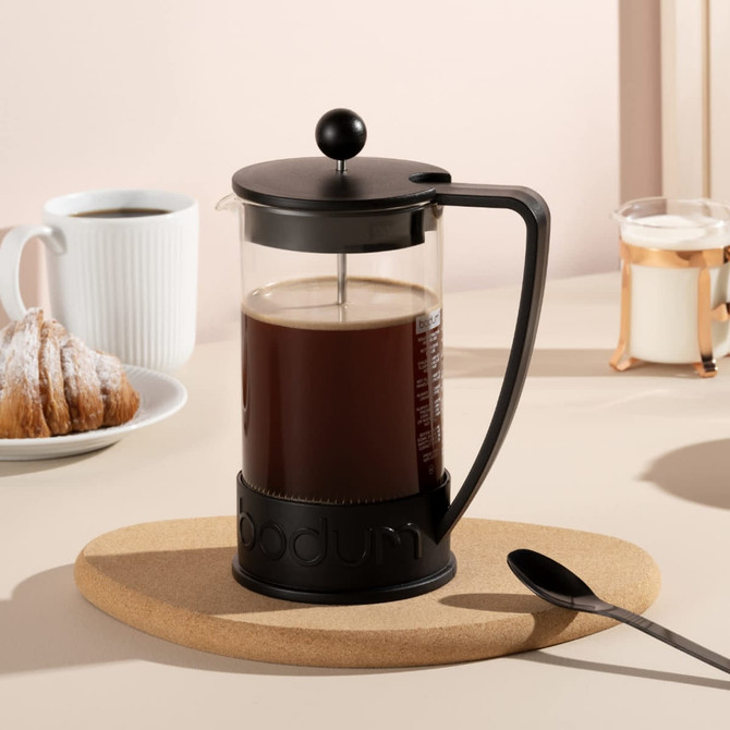 A Bodum Brazil French Press in black, filled with coffee, set on a cork mat, with a spoon and pastries in the background.