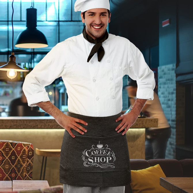 A man wearing a white chef's coat and a charcoal grey apron with a logo stands in a cafe setting.