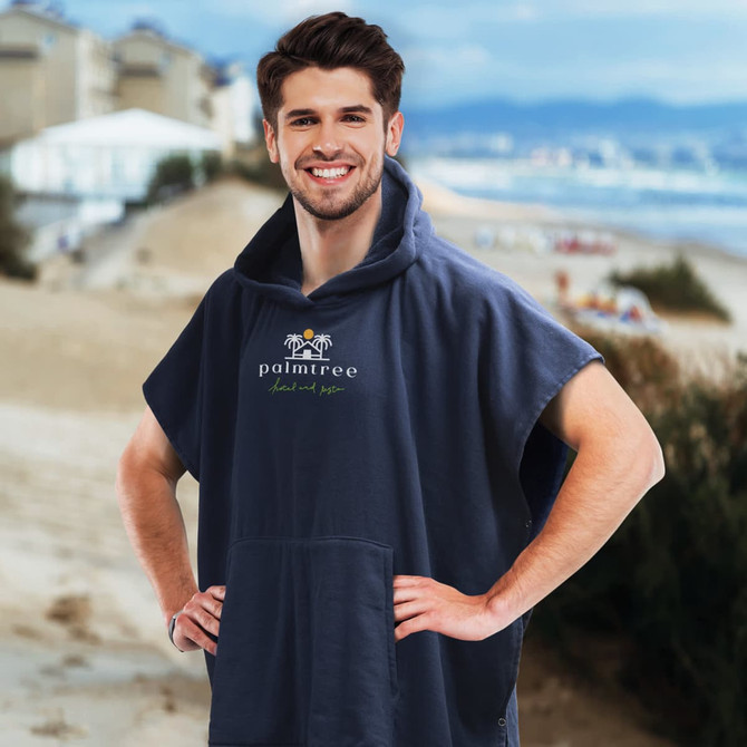 A navy hooded towel with a front pocket and a logo, worn by a smiling man on a beach.