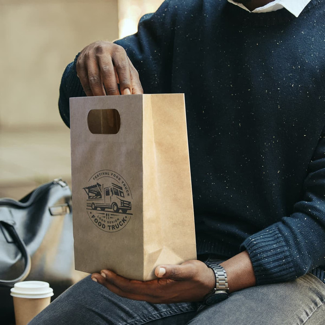 A medium die cut paper bag in brown, featuring a logo, held by a person with a black sweater.