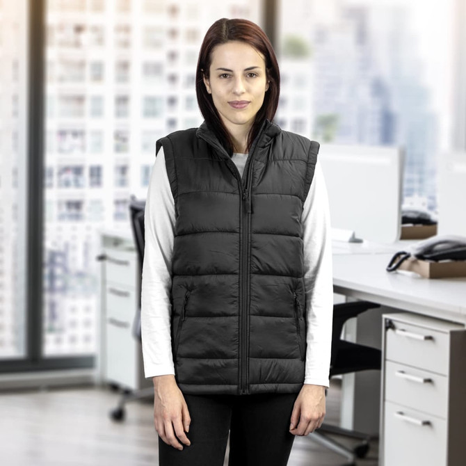 A woman in a black puffer vest stands in an office environment with large windows and modern workspaces.