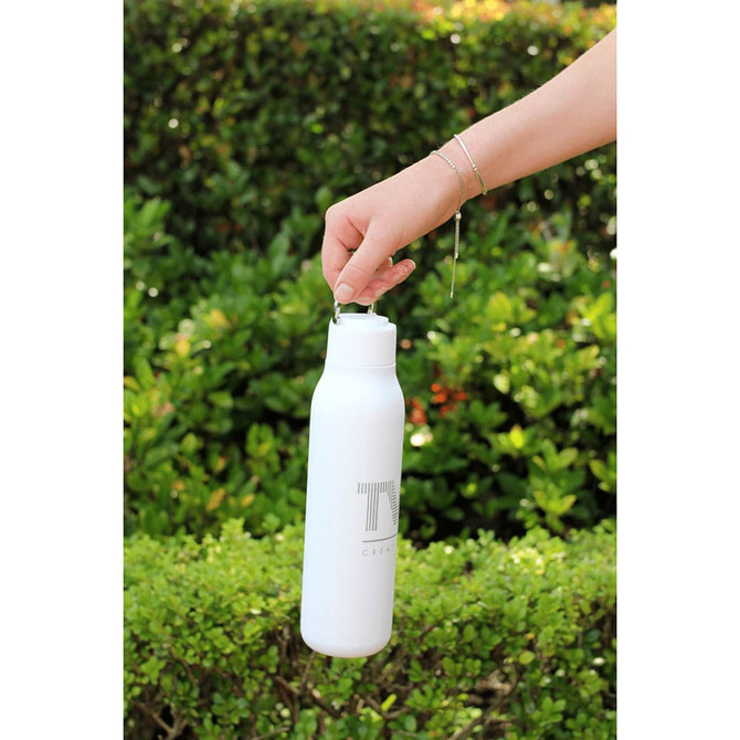 A drink bottle in white with a metal loop, designed for easy carrying, set against greenery.