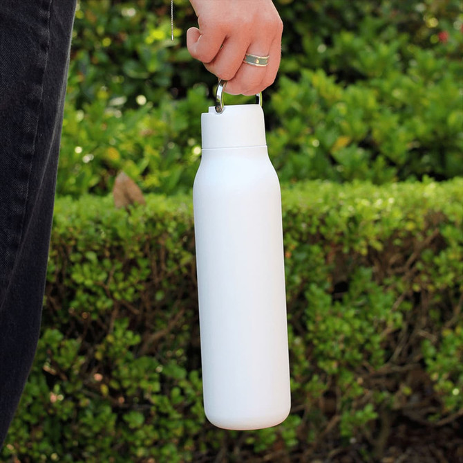 A drink bottle in white with a metal loop, featuring a sleek design, held by a person against a green background.