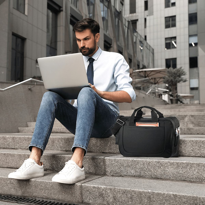 A black laptop briefcase sits on steps beside a man working on a silver laptop, dressed in a white shirt and tie.