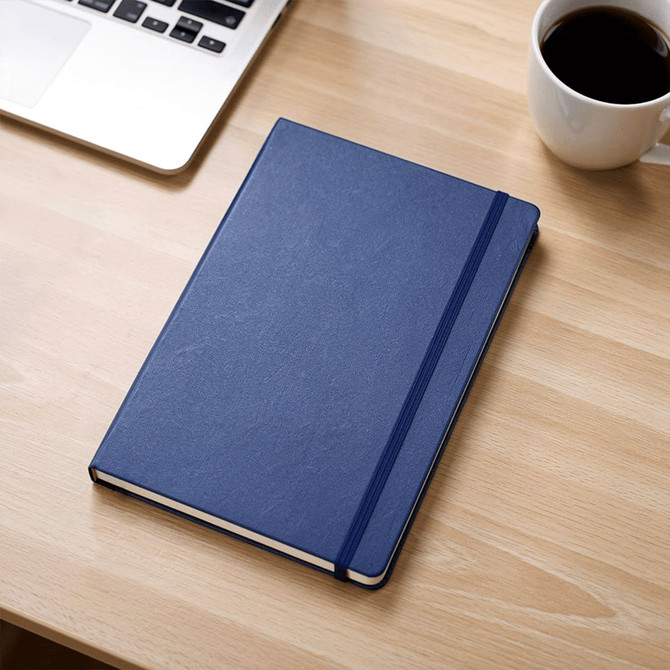 A navy blue journal with a textured cover, placed on a wooden desk next to a laptop and a cup of coffee.