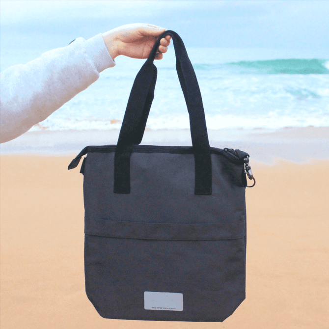 A black laptop tote bag made from recycled material, featuring two handles and a zipper closure, being held by a person.
