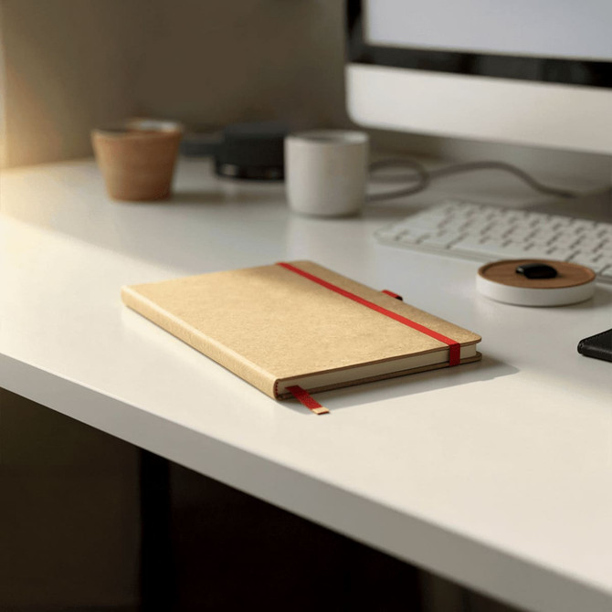 A brown journal with a red elastic band rests on a minimalist desk, accompanied by a cup and electronic devices.