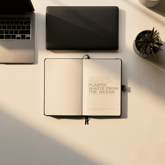 A black journal with a soft cover, open to blank pages, beside a laptop and a small plant.