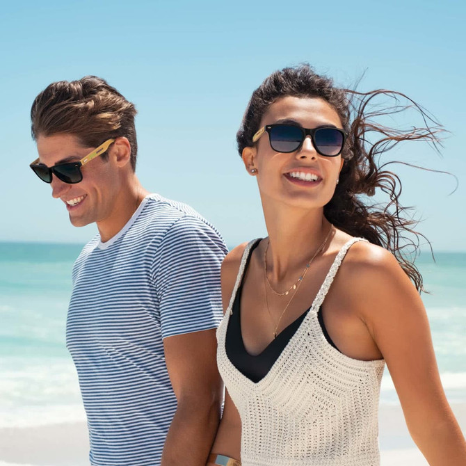 Bamboo sunglasses in a stylish design, worn by a man and woman on a beach, with ocean and blue sky in the background.