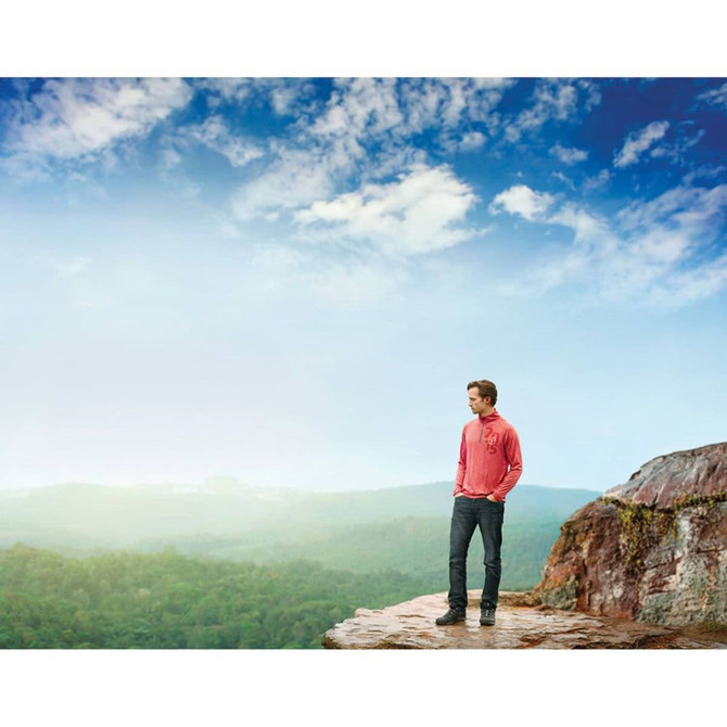 A man wearing a red quarter zip jumper stands on a rocky outcrop, overlooking a lush green landscape under a blue sky.