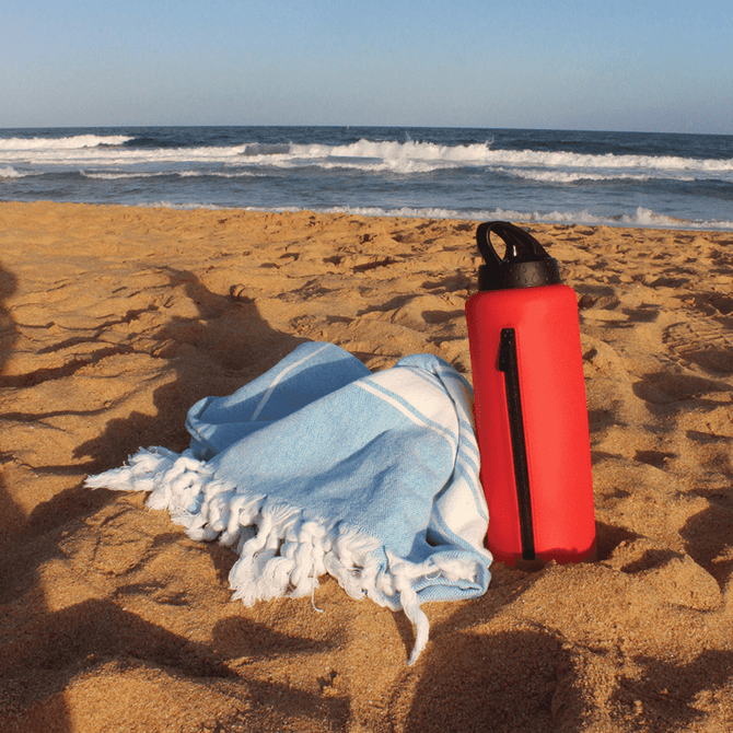 A drink bottle in red, placed on the sand next to a light blue towel, with ocean waves in the background.