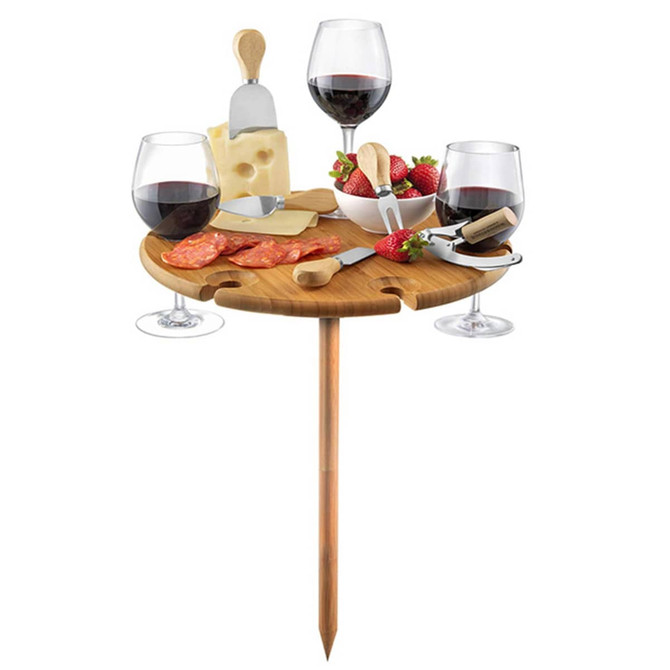 A wooden outdoor party table with a stake, holding wine glasses, cheese, strawberries, and utensils.