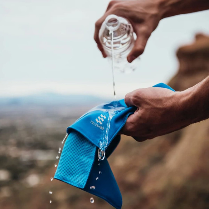 A blue cooling towel is being drenched with water, held in a hand against a scenic outdoor backdrop. It features a logo.