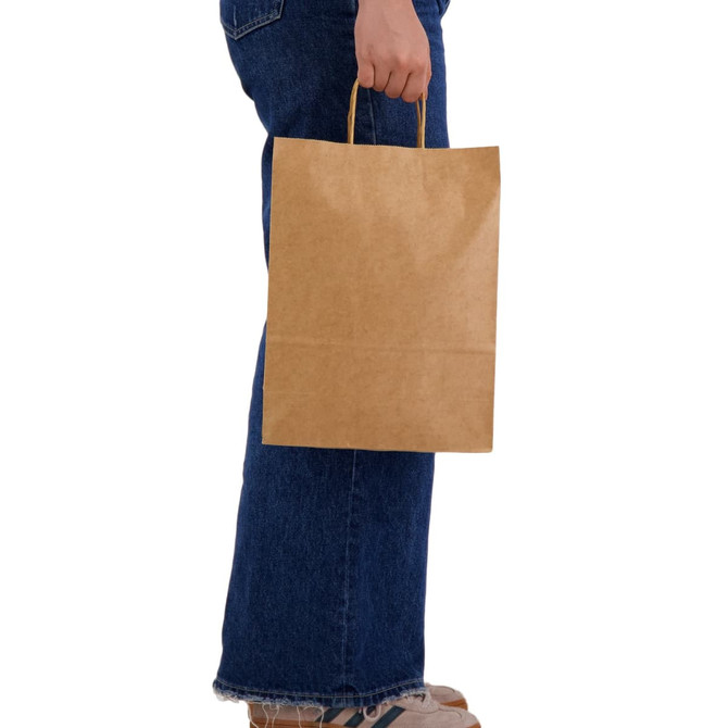 A large brown paper bag held by a person wearing blue jeans and casual shoes. The bag has handles and a simple design.
