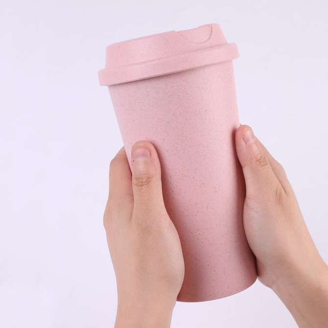 A reusable coffee cup in soft pink, being held in two hands, featuring a lid and a textured finish.