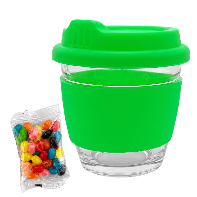 A reusable coffee cup with a clear glass body and a bright green silicone lid and sleeve, alongside a packet of colourful jelly beans.