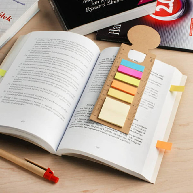 A bookmark ruler sticky note pad with various coloured sticky notes is placed on an open book, alongside a pen and books.
