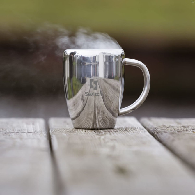 A silver coffee mug with a reflective surface and steam rising, placed on a wooden table. It features a logo.