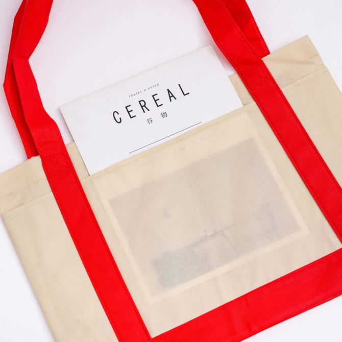 A beige reusable grocery bag with red handles features a clear pocket, holding a magazine inside.