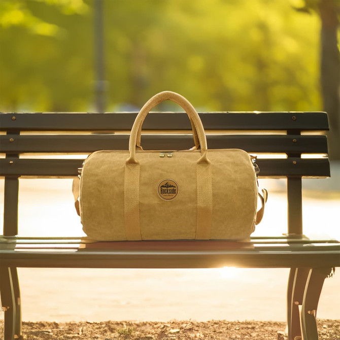 A kraft paper duffle bag in a tan colour sits on a park bench, featuring handles and a logo.