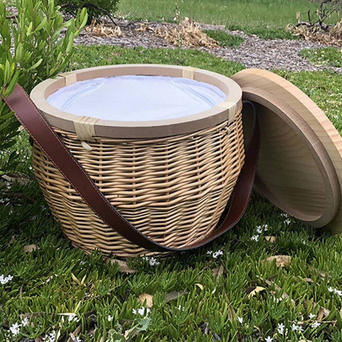 A round wicker picnic cooler basket in natural tones, featuring a wooden lid and a shoulder strap.