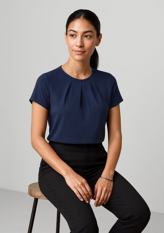 A navy women's short sleeve top with a pleated detail, displayed on a model sitting on a stool.