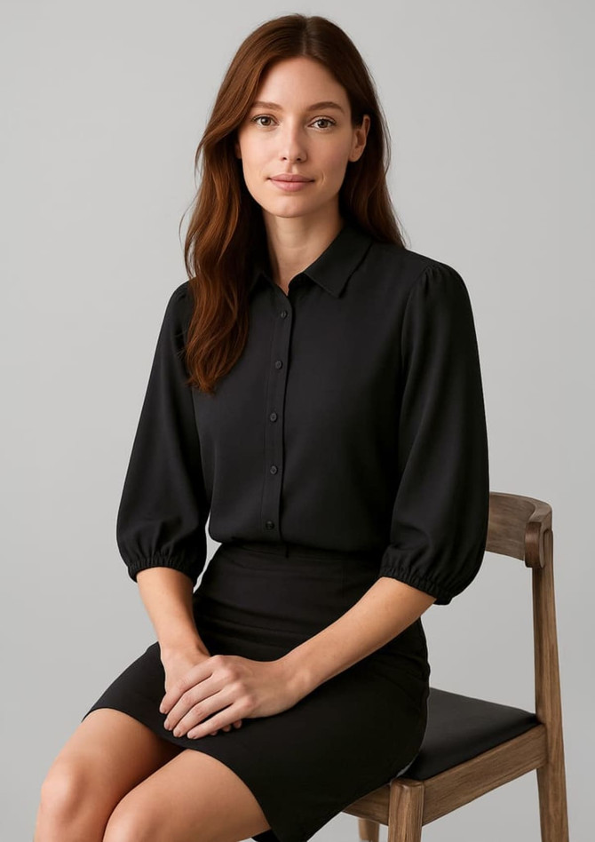A black 3/4 sleeve blouse with a collared neckline, styled with a fitted skirt, displayed on a seated model.