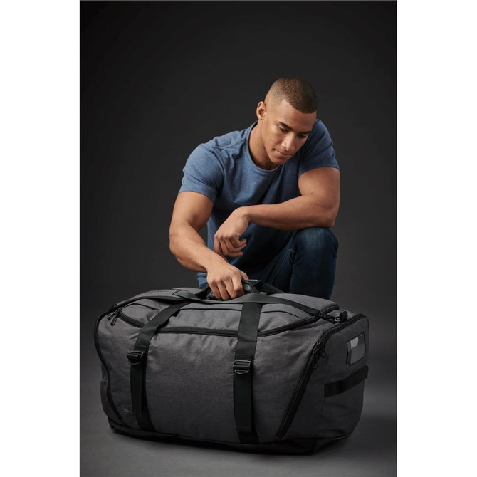A grey duffle bag with green straps sits on the floor next to a man. The bag has a logo and a zippered compartment.