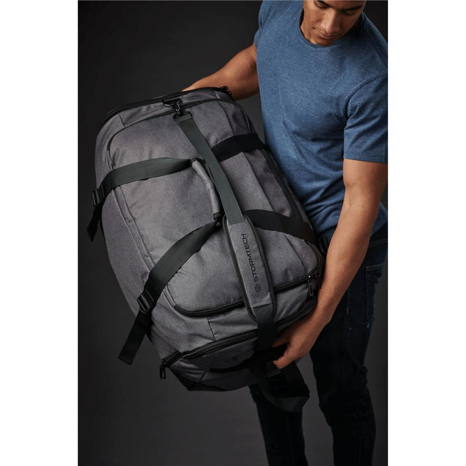 A grey duffle bag with green straps being lifted by a person. The bag has a logo on it.