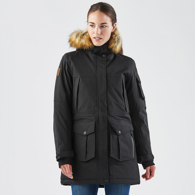 A black women's parka jacket featuring a fur-lined hood and multiple pockets. The model is wearing it in a neutral setting.