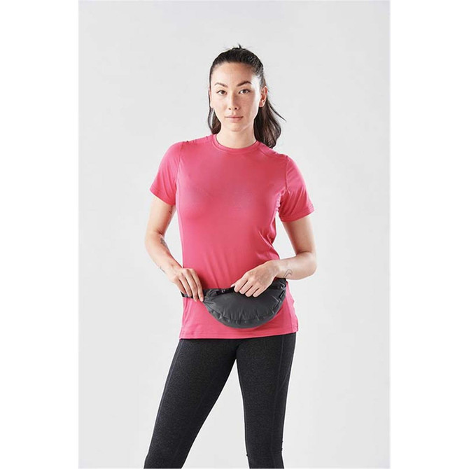 A woman in a pink t-shirt holds a black waist pack, standing against a plain background.