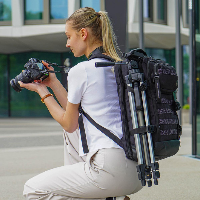 A woman with a black Swissdigital Berg XL Backpack crouches, holding a camera. The backpack has a tripod attached.