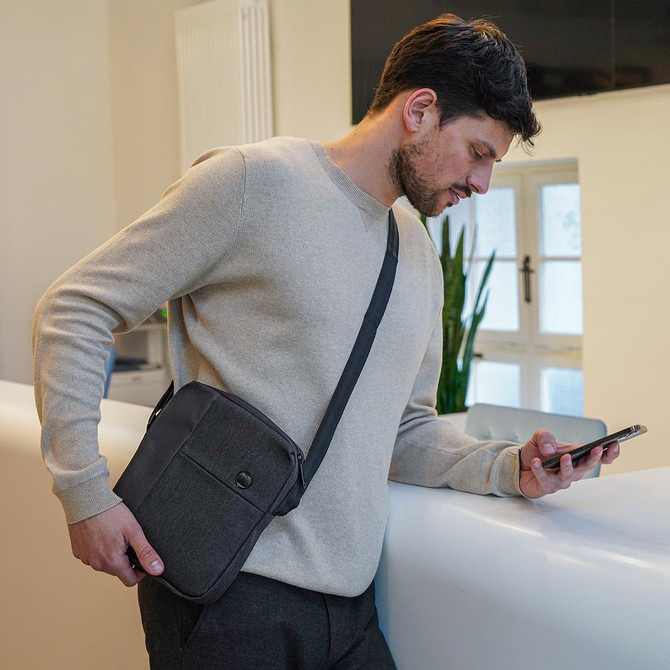 A shoulder bag in grey with a shoulder strap, held by a man in a light sweater, looking at his phone.