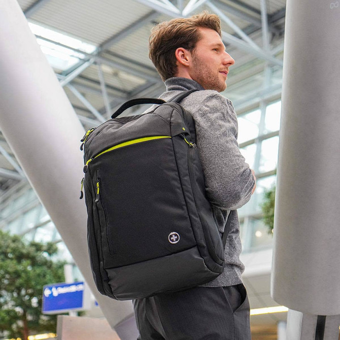 A black backpack with green accents is worn by a man in an indoor setting. The bag features multiple pockets and a logo.